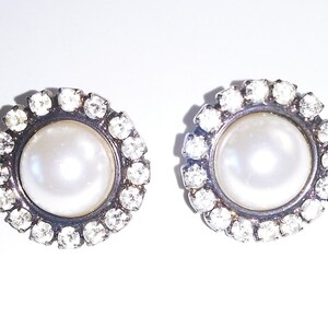 May include: A pair of silver earrings with a white pearl center surrounded by a circle of clear rhinestones.