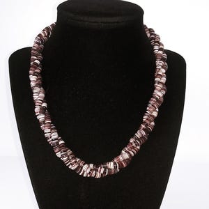 May include: A braided necklace displayed on a black mannequin. The necklace is composed of tightly wound strands in shades of brown, white, and a darker maroon. The design is a twisted rope style, creating a textured and visually interesting accessory.