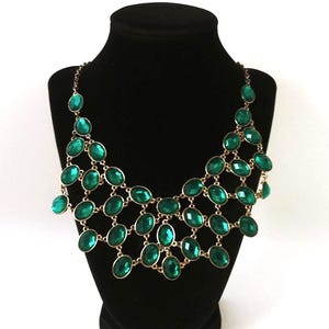 May include: A statement necklace featuring emerald green oval-shaped gemstones set in a gold-tone metal. The necklace is displayed on a black mannequin, showcasing its intricate design and vibrant color. The gemstones are arranged in a cascading pattern.