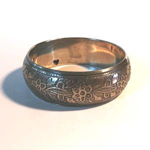 May include: A silver-toned bangle bracelet with an engraved floral design. The bracelet features detailed carvings of flowers and leaves, with a small heart-shaped cutout. The bracelet is approximately 1 inch wide.