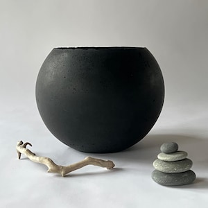 以下が含まれることがあります： 滑らかでわずかにざらざらした表面を持つ、黒い丸いコンクリート製のプランター。手前に小さな枝と灰色の石の積み重ねがある。