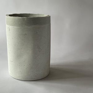 May include: A cylindrical, off-white concrete vase with horizontal ribbed detailing. The top rim and a band near the top are a darker gray. The vase has a rough, textured surface and is set against a plain white background.