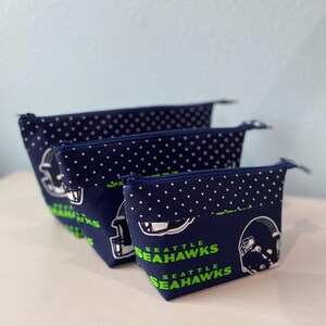 May include: Three navy blue zippered pouches with white polka dots and the words "SEATTLE SEAHAWKS" in green. The pouches feature a football helmet graphic. The pouches are arranged in a row, decreasing in size.