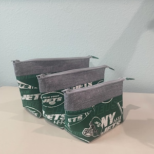 May include: Three zippered pouches with a green and white New York Jets football team design. The top portion of each pouch is gray. The pouches are arranged in a row, with the largest in the back and the smallest in the front.