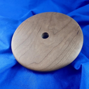 May include: A round, wooden object with a central hole. The wood has a light brown color with visible grain patterns. The object appears to be a lid or a decorative piece.