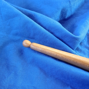 May include: A close-up of a wooden drumstick with a rounded tip. The drumstick is lying on a blue fabric surface.