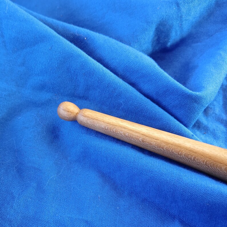 May include: A close-up of a wooden drumstick with a rounded tip. The drumstick is lying on a blue fabric surface.