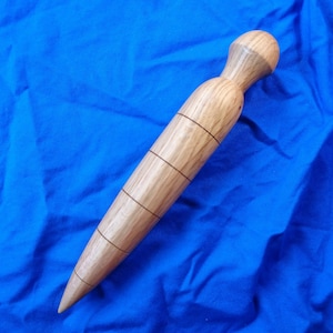 May include: A wooden massage tool with a rounded handle and a tapered end. The tool is light brown with darker lines etched around the body. The tool is lying on a blue fabric surface.