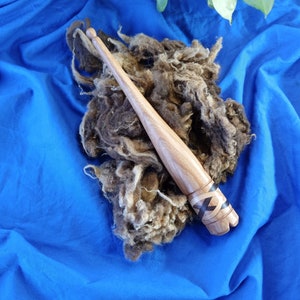 May include: A wooden spinning spindle with a dark wood body and a black and white inlay design. The spindle is lying on a blue fabric surface with brown wool fibres.