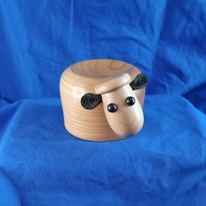 May include: A wooden sheep figurine with black felt ears and eyes. The sheep is lying down with its head turned to the side.