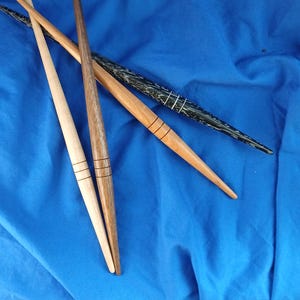 Puede incluir: Cuatro agujas de tejer de madera en varios tonos de marrón y negro, dispuestas en diagonal sobre un fondo azul. Las agujas tienen líneas decorativas y terminan en punta. Se aprecia la veta de la madera.