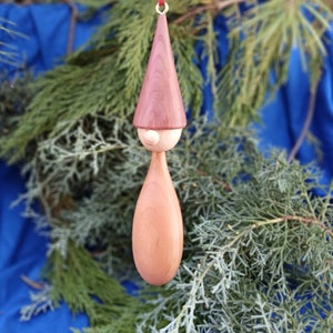 May include: A wooden gnome ornament with a red hat and a light brown body. The gnome is hanging from a red ribbon.