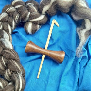 May include: A wooden spindle and a crochet hook rest on a blue fabric, surrounded by brown and white wool roving and a braided yarn. The spindle is dark brown, and the hook is light wood. The yarn and roving are for fiber art.