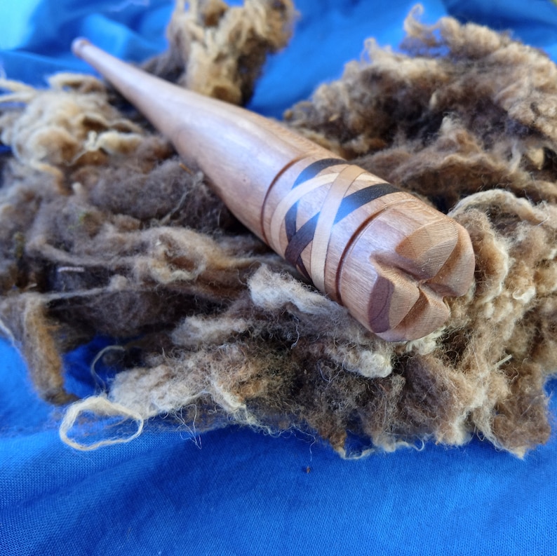 May include: A wooden dowel with a carved design and black and brown inlay. The dowel is resting on a pile of brown wool fibres.
