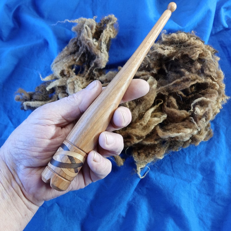 May include: A wooden spinning spindle with a decorative inlay of dark wood and light wood. The spindle is held in a hand, and a pile of brown wool is in the background.