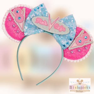 May include: A whimsical headband featuring pink sequined ears with a white trim and a light blue sequined bow. The headband has a light blue band and a gray emblem with the text "Flo's Cafe".