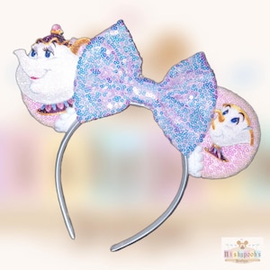 May include: A headband featuring two circular ears decorated with glittery pink and white character appliques of a teapot and a teacup. A large, central bow covered in iridescent blue and purple sequins sits between the two ears.