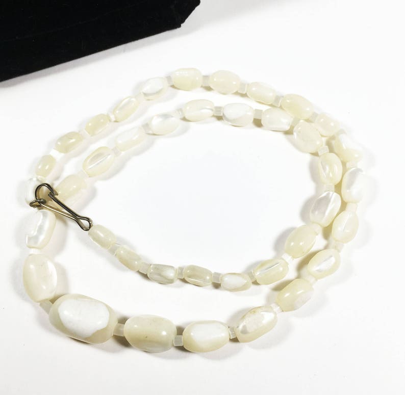 May include: A white pearl necklace with a gold clasp. The pearls are oval shaped and have a slight iridescent sheen.