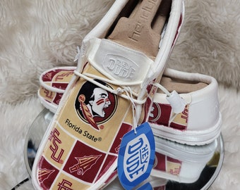 May include: A pair of white canvas slip-on shoes with a Florida State Seminoles design. The shoes have a red, gold, and white pattern with the Florida State logo and the words "Florida State" on the side. The shoes are on a white fuzzy surface and a silver round surface. The shoes have a blue tag that says "Hey Dude" on it.