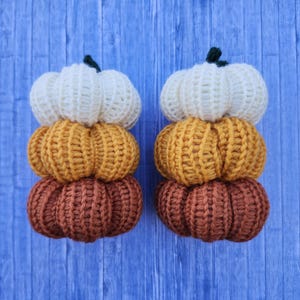 May include: Two stacks of three crocheted pumpkins in shades of white, yellow-orange, and brown. Each pumpkin has a ribbed texture and a green stem. The pumpkins are arranged on a blue wooden surface.