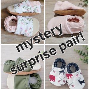 May include: A collage of baby shoes in various styles and colors. The shoes include floral prints, solid pink, and a pair with a bear and arrow design. The text "mystery surprise pair!" is overlaid on the image.