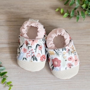 May include: A pair of baby shoes with a floral print. The shoes are made of soft fabric and have a light pink lining. The shoes have a beige sole and the text "Tiny Toes" is printed on the top of one shoe.