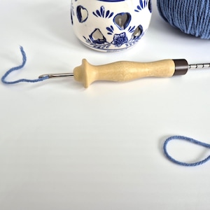 Könnte beinhalten: Ein hölzernes Punch-Needle-Werkzeug mit einer Metallnadel und einer braunen Spitze, neben einer blauen Garnschlaufe. Ein blau-weißer Keramik-Kerzenhalter und ein Knäuel blauen Garns befinden sich im Hintergrund. Werkzeuge zum Teppichhaken und andere Handarbeiten.