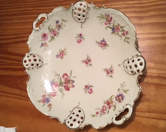 May include: A white porcelain serving platter with a delicate floral pattern in shades of pink, purple, and green. The platter has a scalloped edge and two openwork handles.