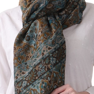 May include: A teal, brown, and gold paisley patterned scarf with fringed edges. The scarf is wrapped around a person's neck and tied in a knot at the front.