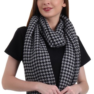 May include: A black and gray houndstooth patterned scarf. The scarf is folded and draped around the neck of a person wearing a black t-shirt and blue jeans.