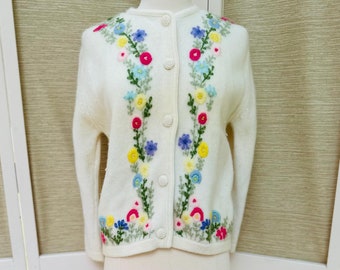 May include: A white cardigan sweater with colorful floral embroidery. The cardigan has a button-up front and long sleeves.