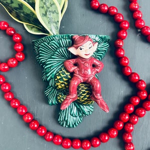 Op de afbeelding: Een groene keramische plantenpot in de vorm van een dennenappel met een rood elfje dat er bovenop zit. De pot is versierd met gele dennenappels en groene bladeren. Een streng rode kralen is om de pot gewikkeld.