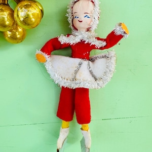 Op de afbeelding: Een vintage kerstversiering met een meisje dat een rode jurk draagt met witte bontkraag en schaatsen. Ze heeft een geschilderd gezicht met blauwe ogen en een roze teint.