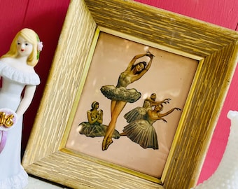May include: A framed print of a vintage ballet performance featuring five dancers in white tutus and pink tights. The dancers are posed in a graceful and elegant manner. The frame is gold with a wood grain finish.