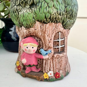 Könnte beinhalten: Ein skurriler Keramik-Pflanztopf in Form eines Baumstamms mit einem grünen, belaubten Oberteil. Ein kleiner Elf in rosa Kleidung hält einen blauen Vogel. Der Pflanztopf hat ein kleines Fenster und ist mit bunten Blumen verziert.
