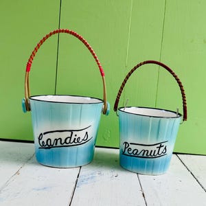 May include: Two light blue and white gradient metal buckets with black rims and handles. The larger bucket features the word "candies" in black script, while the smaller bucket features the word "Peanuts" in black script. Both have red accents.