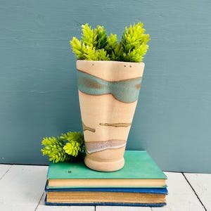 Könnte beinhalten: Eine beige Keramikvase mit türkisfarbenen und braunen Akzenten, die grüne Kunstpflanzen enthält. Die Vase steht auf zwei alten Büchern mit türkisfarbenen Einbänden und goldfarbenen Seiten. Der Hintergrund ist eine türkisfarbene Wand.