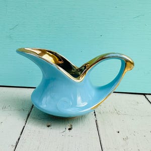 Op de afbeelding: Een lichtblauwe keramische creamer met een gouden rand en handvat. De creamer heeft een gebogen tuit en een decoratief wervelontwerp aan de zijkant. Het handvat en de rand zijn afgewerkt met goud. De creamer is ongeveer 15 cm lang.