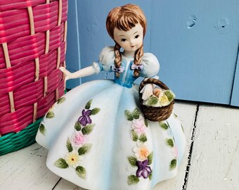 Op de afbeelding: Een porseleinen beeldje van een meisje in een blauwe jurk met bloemenaccenten. Ze houdt een mand met bloemen vast en heeft bruin haar in vlechten.