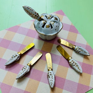 May include: Set of five white ceramic butter knives with gold accents and grape designs. The knives are arranged on a pink and white checked surface with a white ceramic butter dish with gold accents in the centre.