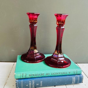May include: Two ruby red glass candle holders with a geometric design. The candle holders are placed on top of two vintage books with teal and blue covers. The candle holders have a flared top and a wide base.