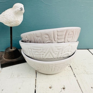 Puede incluir: Tres cuencos de cerámica en tonos blanco y gris apilados sobre una superficie de madera blanca. Cada cuenco presenta un patrón geométrico texturizado. A la izquierda, una estatua de pájaro blanco sobre un soporte de metal negro, sobre un fondo turquesa.