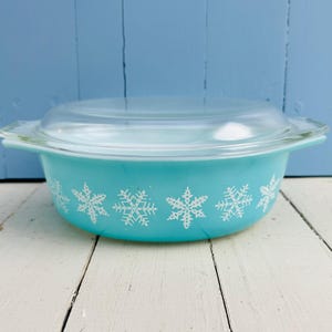 May include: A vintage turquoise Pyrex casserole dish with a clear glass lid. The dish features a white snowflake pattern around the exterior. The dish is on a white wooden surface with a blue background.