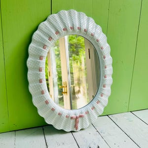 May include: Oval white mirror with a scalloped edge and a dotted texture. The frame features small pink rectangular accents and a pink bow at the bottom. The mirror reflects a bright outdoor scene.