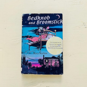 May include: A paperback book titled Bedknob and Broomstick by Mary Norton. The cover features a dark blue night sky, a brass bed frame, and a house silhouette. A circular yellow sticker states it inspired the Walt Disney film.