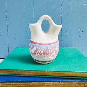 May include: A matte white ceramic vessel featuring a central arched handle and two openings. The lower portion is decorated with horizontal bands in shades of pink and blue, accented by etched geometric patterns that reveal the white base color.