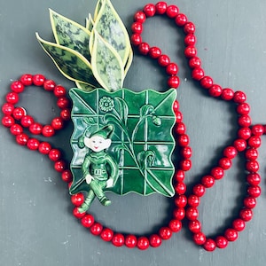 May include: A green ceramic planter shaped like a leaf with a small elf figurine hanging from the side. The planter is filled with a green and white variegated plant. A strand of red beads is wrapped around the planter.