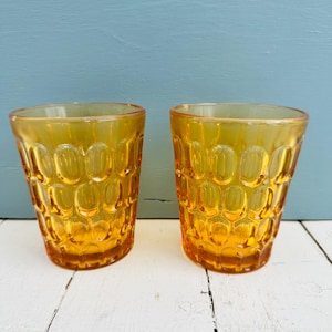 Puede incluir: Dos vasos de color ámbar. Cada vaso tiene un patrón ovalado texturizado en el exterior. Los vasos están hechos de un material transparente. Los vasos están sobre una superficie de madera blanca.