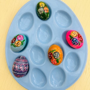 May include: A light blue egg holder with several decorated Easter eggs. The eggs are painted with floral designs in various colors, including pink, green, orange, and blue. The holder has oval indentations to hold the eggs.