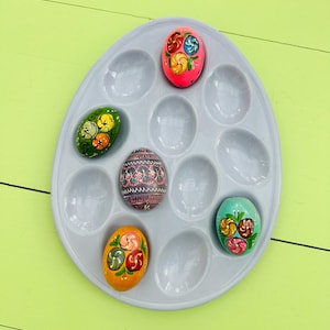 May include: A gray plastic egg tray holding several decorated Easter eggs. The eggs are painted with floral designs in various colors, including pink, yellow, green, and blue. The tray is on a light green surface.